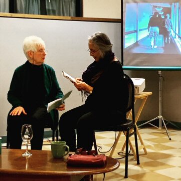 two women are seated behind a table.  a picture of people in a corridor is projected on a screen behind them.